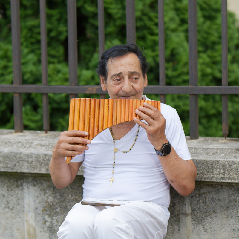 26 - Old man performing panflute music near a stone fence in a public place